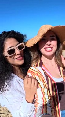 Güneşli bir günde kumlu bir sahilde selfie çekerken gülümseyen ve gülen iki güzel genç kadın. Neşeli bayan arkadaşlar okyanus kıyısındaki yaz tatilinin tadını çıkarıyorlar.