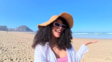 Kıvırcık saçlı, kumsalda video izlerken kameraya konuşan güzel, mutlu bir kadın. Gülümseyen nüfuzlu kişi, deniz kenarında yaz tatilinde canlı yayında takipçilerini selamlıyor.