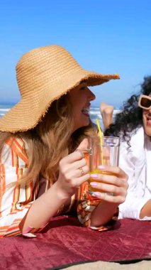 İki güzel, gülümseyen kadın kumlu bir sahilde uzanmış taze portakal suyu içiyor ve açık mavi gökyüzünün altında arkadaşça sohbet ederken yaz tatillerinin tadını çıkarıyorlar.