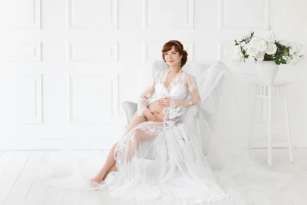 Femme enceinte assise sur une chaise dans une belle robe blanche boudoir (négligée ). — Photo