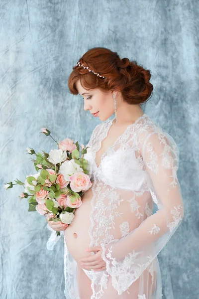 Mulher grávida segurando flores, de pé no vestido boudoir (negligee) em um fundo azul . — Fotografia de Stock