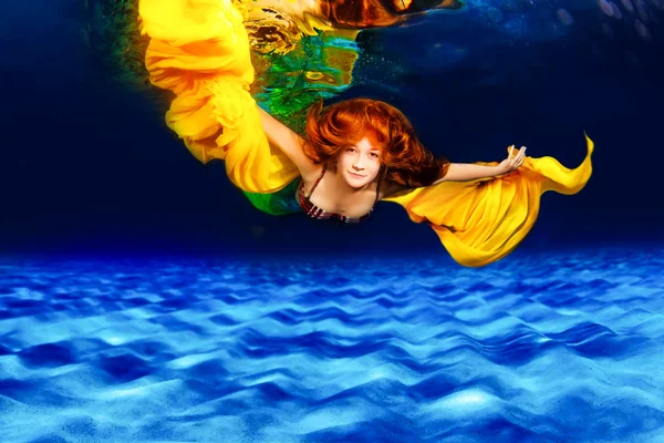 Piękna rudowłosa dziewczyna pływająca pod wodą w morzu. Pani w trzepoczącym jedwabiu piękna sukienka, swinging tkanina w wodzie, piękna dziewczyna z długim latający żółty materiał. — Zdjęcie stockowe