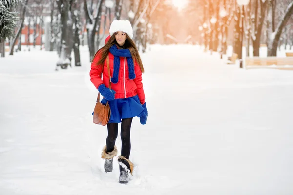 在白色的皮毛帽子，一件红色的外套和蓝色手套，一个女孩在公园散步在冬天. — 图库照片