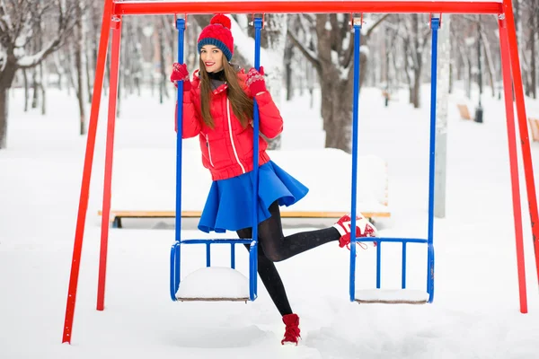 Hermoso invierno Retrato de una mujer joven en invierno paisaje cubierto de nieve. Una chica con una ropa azul brillante y roja de pie cerca de los columpios . — Foto de Stock