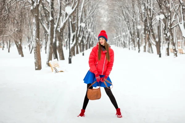 美丽的冬天冬天下雪的景色的年轻女子的画像。一顶红色帽子，红夹克，蓝色的裙子，女孩在公园里摆在冬天. — 图库照片