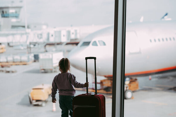 Маленькая девочка в аэропорту в маске для лица держит ручную кладь. Ребенок смотрит на самолет после вспышки коронавируса. Безопасное путешествие и полет с ребенком в условиях вирусной пандемии. Парень у выхода. Отпуск после блокировки ковида-19.