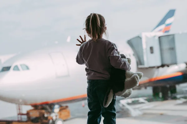 stock image Toddler girl at airport in face mask holding a toy. Child look at airplane after coronavirus outbreak. Safe travel and flying with child in virus pandemic. Kid at departure gate. Vacation after covid-19 lockdown.