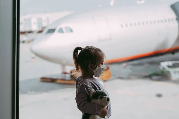 おもちゃを保持顔マスクの空港で幼児の女の子 子供はコロナウイルス発生後飛行機を見る 安全な旅行とウイルスのパンデミックで子供と一緒に飛ぶ 出発ゲートで子供 Covid 19ロックダウン後の休暇 ストック画像