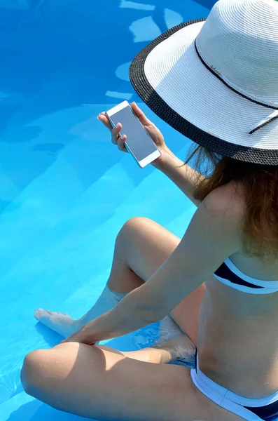Una mujer con sombrero y un smartphone en la mano, foto junto a la piscina . — Foto de Stock