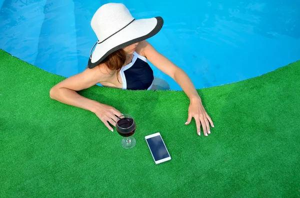 Mujer joven con sombrero cerca de la piscina, con vino y un teléfono inteligente — Foto de Stock