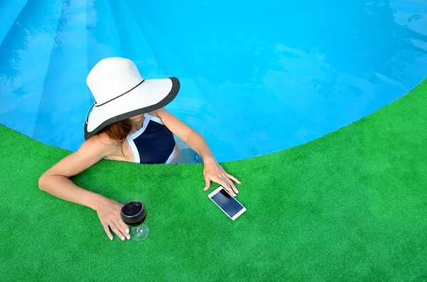 Mujer joven con sombrero cerca de la piscina, con vino y un teléfono inteligente — Foto de Stock