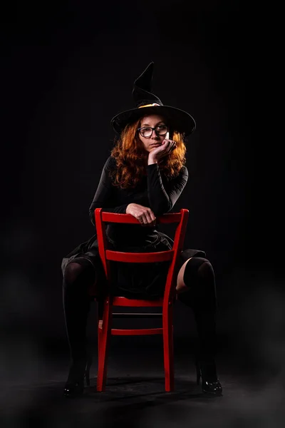Siyah Elbiseli Siyah Arka Planda Kırmızı Sandalyede Poz Veren Cadı — Stok fotoğraf