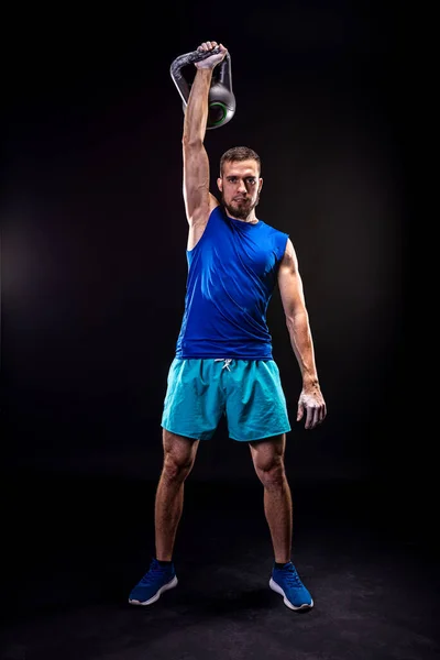 A sports man in a blue T-shirt and shorts trains with a kettlebell on a black background. Fitness with kettlebell. Weight training with a kettlebell.