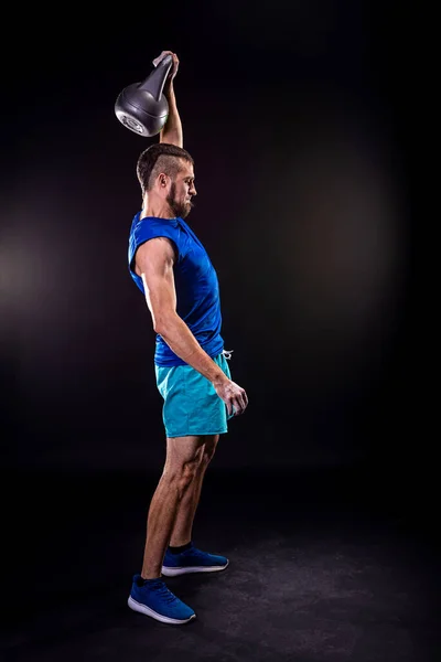 A sports man in a blue T-shirt and shorts trains with a kettlebell on a black background. Fitness with kettlebell. Weight training with a kettlebell.