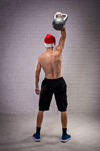 Sports Man Santa Hat Lifts Kettlebell Rear View Fitness Kettlebell — Stock Photo, Image