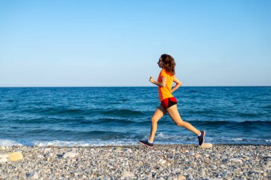Açık turuncu tişörtlü ince bir kadın plaj boyunca koşuyor..