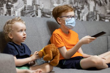 Maskeli çocuklar kanepede oturup televizyon seyrediyor, ağabeyi uzaktan kumandayı elinde tutarak kanalı değiştiriyor. Karantinaya alındı.