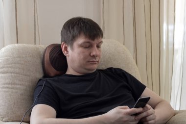 Sandalyede oturan bir adam masaj yastığıyla boynuna masaj yapar ve telefona bakar, mesajlaşır..