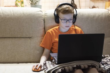 Gözlüklü bir çocuk kulaklıkla müzik dinler ve dizüstü bilgisayarda oturur..