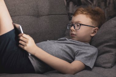 Kanepede bir genç tabletle oynuyor. Gözlüklü okul çocuğu..