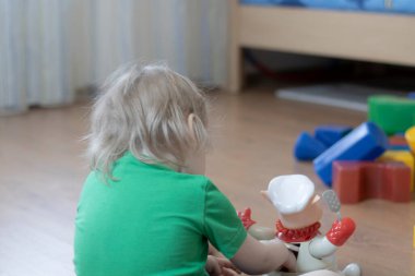 Oturan küçük bir çocuk, arka taraftaki bir oyuncakla çocuk odasında oynuyor..