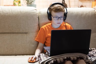 Gözlüklü bir çocuk kulaklıkla müzik dinler ve dizüstü bilgisayarda oturur..