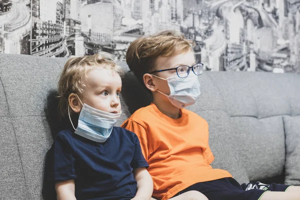 İki kardeş kanepede oturmuş, maske takıp televizyon izliyorlar..