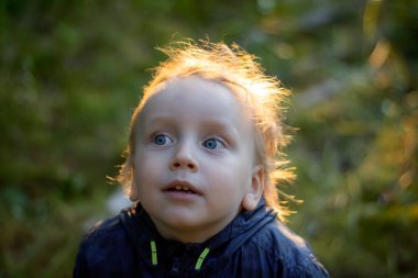 Ceketli, mavi gözlü 3 yaşında bir çocuk yana bakıyor, düşünceli bir bakış, güneş parlıyor. Yakın plan portre.
