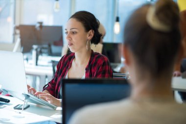 Çok ırklı iş arkadaşları, sarışın, beyaz, latin, esmer, yakışıklı iş kadınları (çalışanlar), modern bir şirkette dizüstü bilgisayar üzerinde çalışırken sarı bir koltukta oturuyorlar.