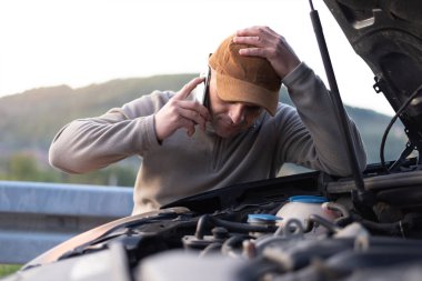 Kızgın genç adam yolun kenarındaki bozuk arabanın yanında otururken yardım bekliyor. Yol kenarı yardım kavramı