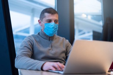 Karantina sırasında maske takan bir iş adamı. Bir kafede genç bir adam pencerenin yanındaki dizüstü bilgisayarda çalışıyor. İzolasyon, koronavirüs, evde çalışmak..