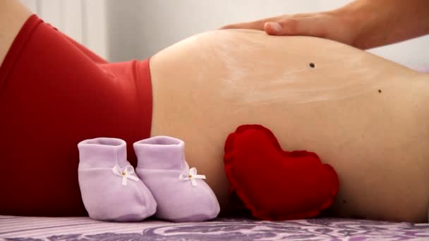 Junge Schwangere und ihr Mann mit Kosmetikcreme sitzen auf einem Sofa im Zimmer. Konzept der Körperpflege — Stockvideo