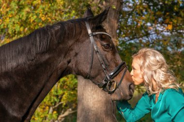 Yeşil elbiseli çekici sarışın kadın kahverengi atı burnundan öpüyor. Sonbaharda ormanda altın renginde.