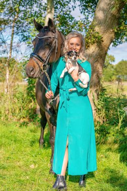 Ormanda atı ve köpeğiyle gülümseyen güzel sarışın kız. Yeşil bir elbise giyiyor. Seçici odak.