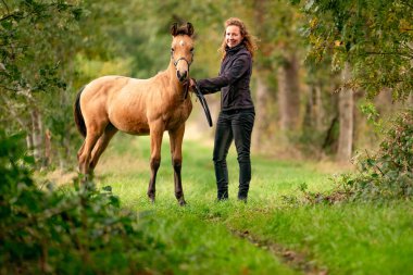 Açık kahverengi bir tayın kadın sahibi, sonbahar güneşi aygırının yanında duruyor..