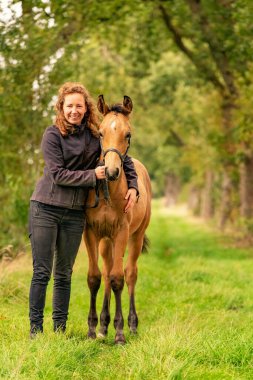 Açık kahverengi bir tayın kadın sahibi, sonbahar güneşi aygırının yanında duruyor..
