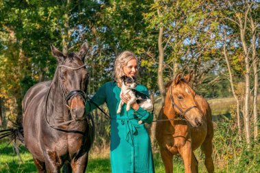 Ormanda atıyla, yavrusuyla ve köpeğiyle gülümseyen sarışın bir kızın güzel portresi. Yeşil bir elbise giyiyor..
