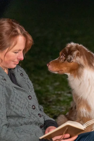 Australian Shepherd cão e mulher na fogueira, A mulher está lendo um livro, sentado em roupas casuais de inverno, Foco no cão, foco seletivo — Fotografia de Stock