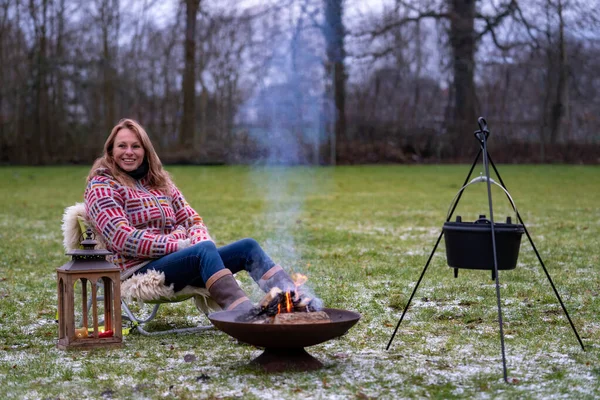 Young blond woman in a wool sweater sits outside on the grass by the campfire. Burning fire in fire bowl a boiling kettle hangs above it, in the late afternoon