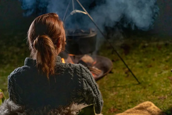 Young blond woman sits outside by the campfire in the woods. It is dark in winter and she is warm by the fire in the forest. Head seen from behind