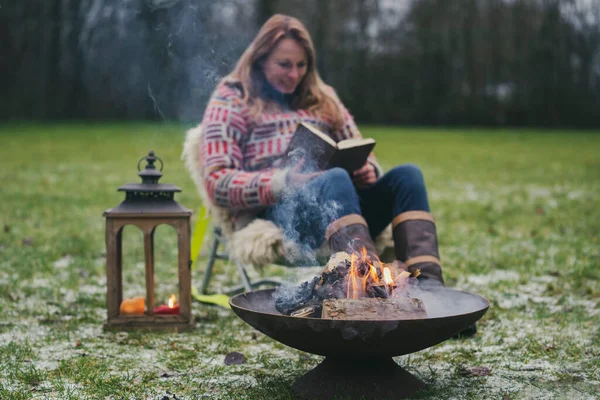 Young blond woman in a wool sweater reads a book outside on the grass by the campfire. Burning fire in fire bowl, a boiling kettle hangs above. camping in winter in the late afternoon, at sunset