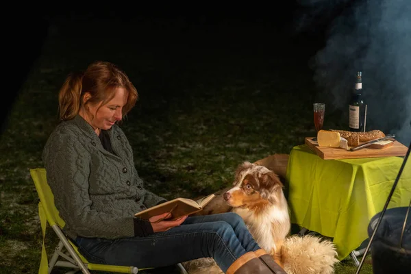 Australian Shepherd dog is lying by the campfire, the kettle is cooking and steaming, The woman is reading a book, sitting in casual winter clothes on a camping chair. cheese and wine are on a table