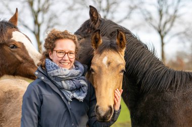 Çayırda bir yaşındaki aygırıyla balmumu montlu genç bir kadın. Sosyalleşmek için onlara sarılıyor. Üç at kafası.