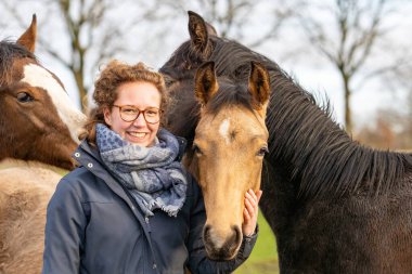 Çayırda bir yaşındaki aygırıyla balmumu montlu gülümseyen genç bir kadın. Sosyalleşmek için onlara sarılıyor. Üç at kafası.