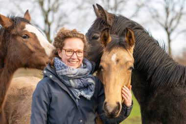 Çayırda bir yaşındaki aygırıyla balmumu montlu gülümseyen genç bir kadın. Sosyalleşmek için onlara sarılıyor. Üç at kafası.