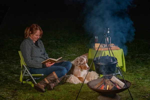 Australian Shepherd dog is lying by the campfire, the kettle is cooking and steaming, The woman is reading a book, sitting in casual winter clothes on a camping chair. cheese and wine are on a table