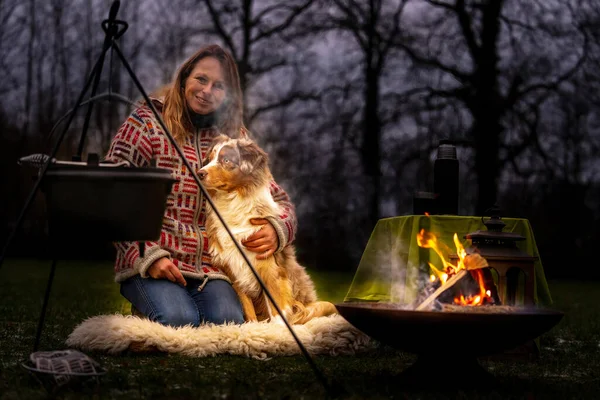 Tricolor Australian Shepherd sits next to a young woman by the campfire. In winter, snow on the grass. The fire is burning at night. Hugging the dog. Selective focus