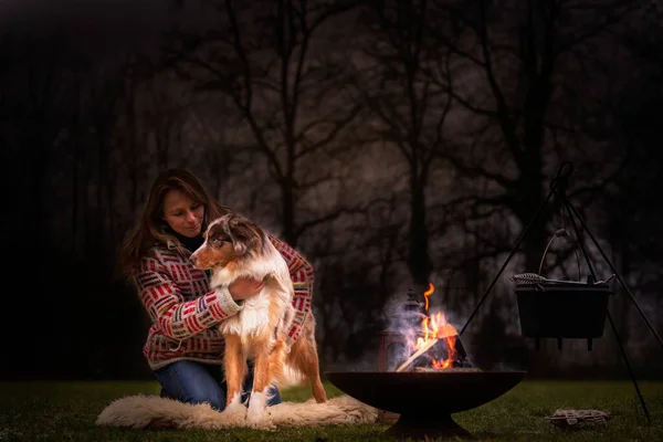 Tricolor Australian Shepherd kamp ateşinin yanında genç bir kadının yanında oturuyor. Kışın, çimlerin üzerinde kar. Ateş geceleri yakıyor. Köpeğe sarılıyorum. Seçici odak