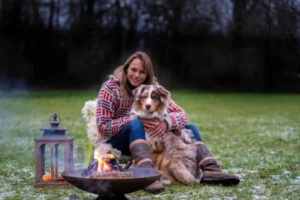 Un pastor australiano se sienta junto a la fogata con su dueño por la tarde. Las velas arden en una linterna. Mujer y perro acurrucarse y disfrutar de la vida de camping, en invierno — Foto de Stock