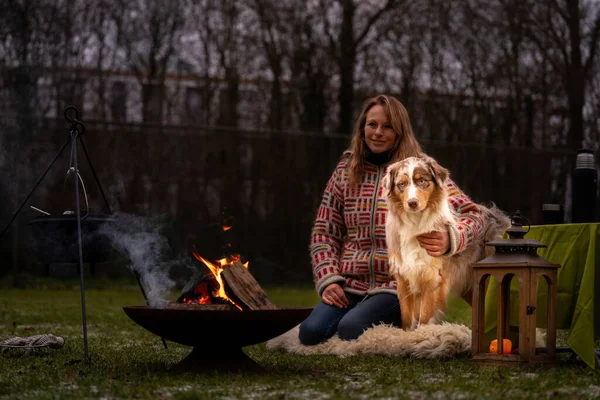 Tricolor pastor australiano se sienta al lado de una joven junto a la fogata. En invierno, nieve en la hierba. El fuego arde por la noche. Abrazar al perro. Enfoque selectivo — Foto de Stock